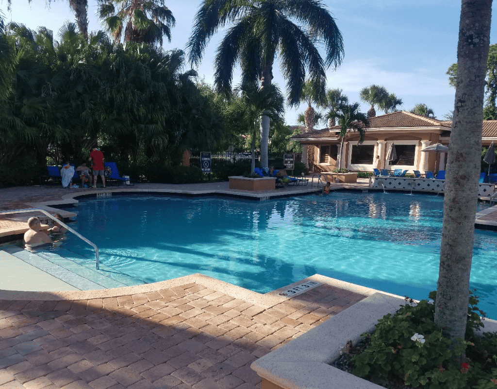 Large resort-style community pool with palm-lined deck and lounge chairs under blue sky, serviced by Pools USA in Port St. Lucie, FL.
